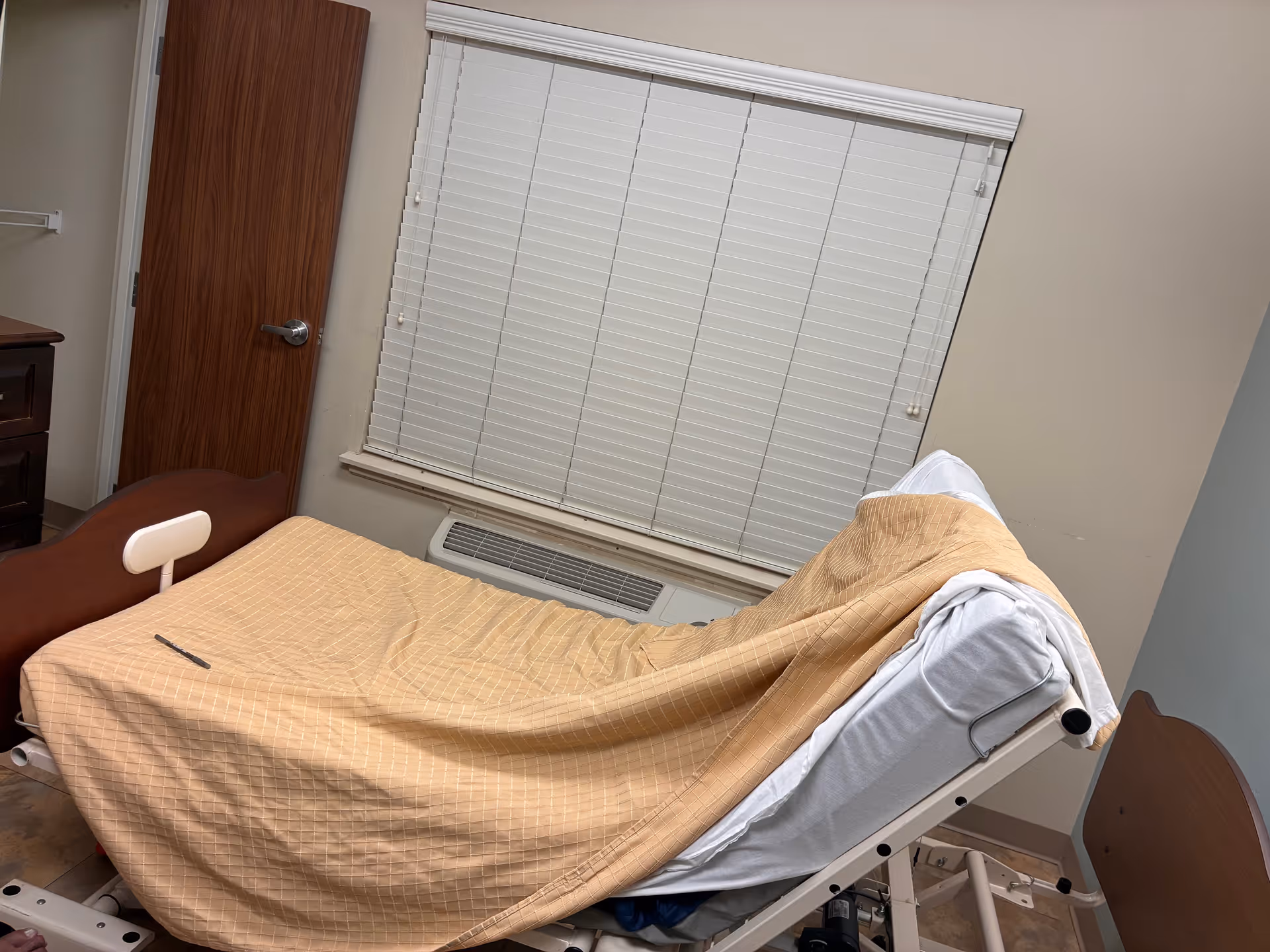 Hospital-style bed in a small patient room with blinds and a beige blanket.