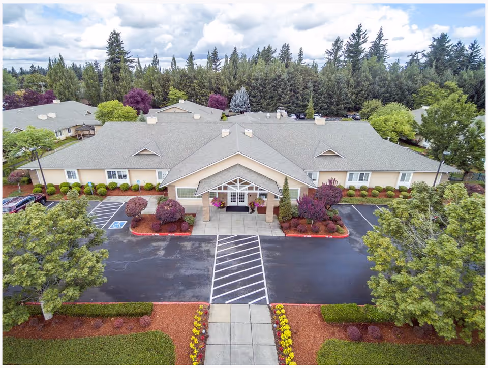 Aerial view of the single-story senior living building with a covered front entrance, landscaped beds, and a parking area surrounded by trees.