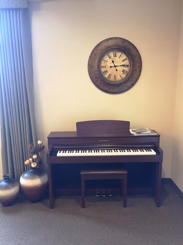 A wooden upright piano with a matching bench is placed against a beige wall. Above the piano hangs a large round wall clock with Roman numerals. To the left of the piano, there are two decorative vases, one taller than the other, with dried floral arrangements. A window with closed curtains is partially visible on the far left side.