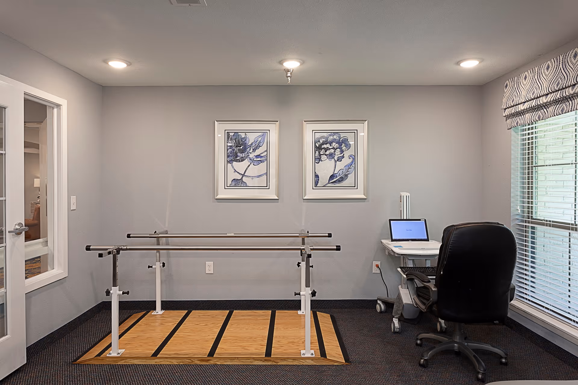 Therapy room with parallel bars, a desk, chair, and framed wall art.