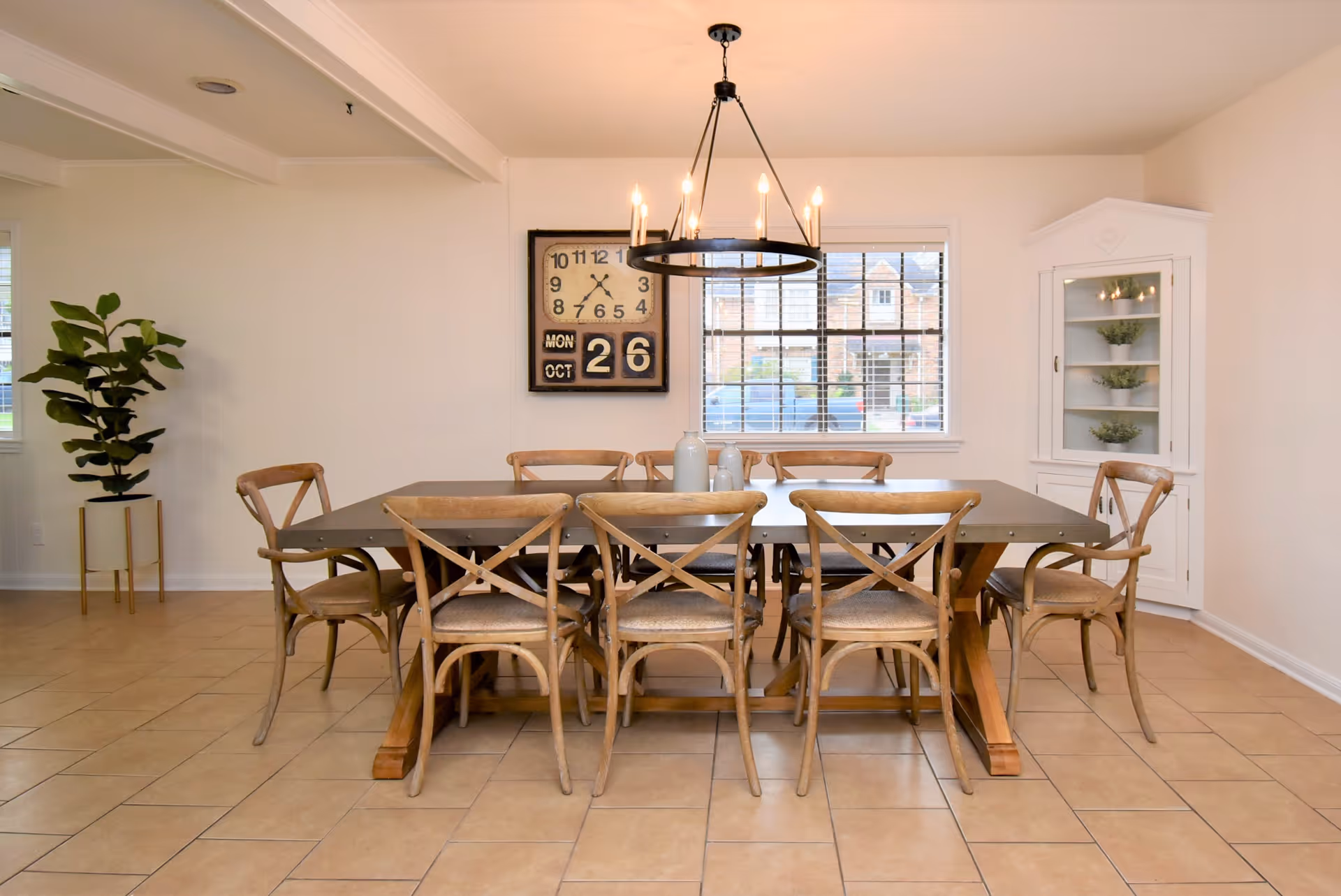 A dining room with a large rectangular wooden table surrounded by eight wooden chairs with woven seats. A modern chandelier with exposed bulbs hangs above the table. On the wall behind the table is a large clock with the date displayed as Monday, October 26. A window with blinds lets in natural light, and a white corner cabinet with glass doors holds small potted plants. A tall potted plant is placed in the corner of the room.