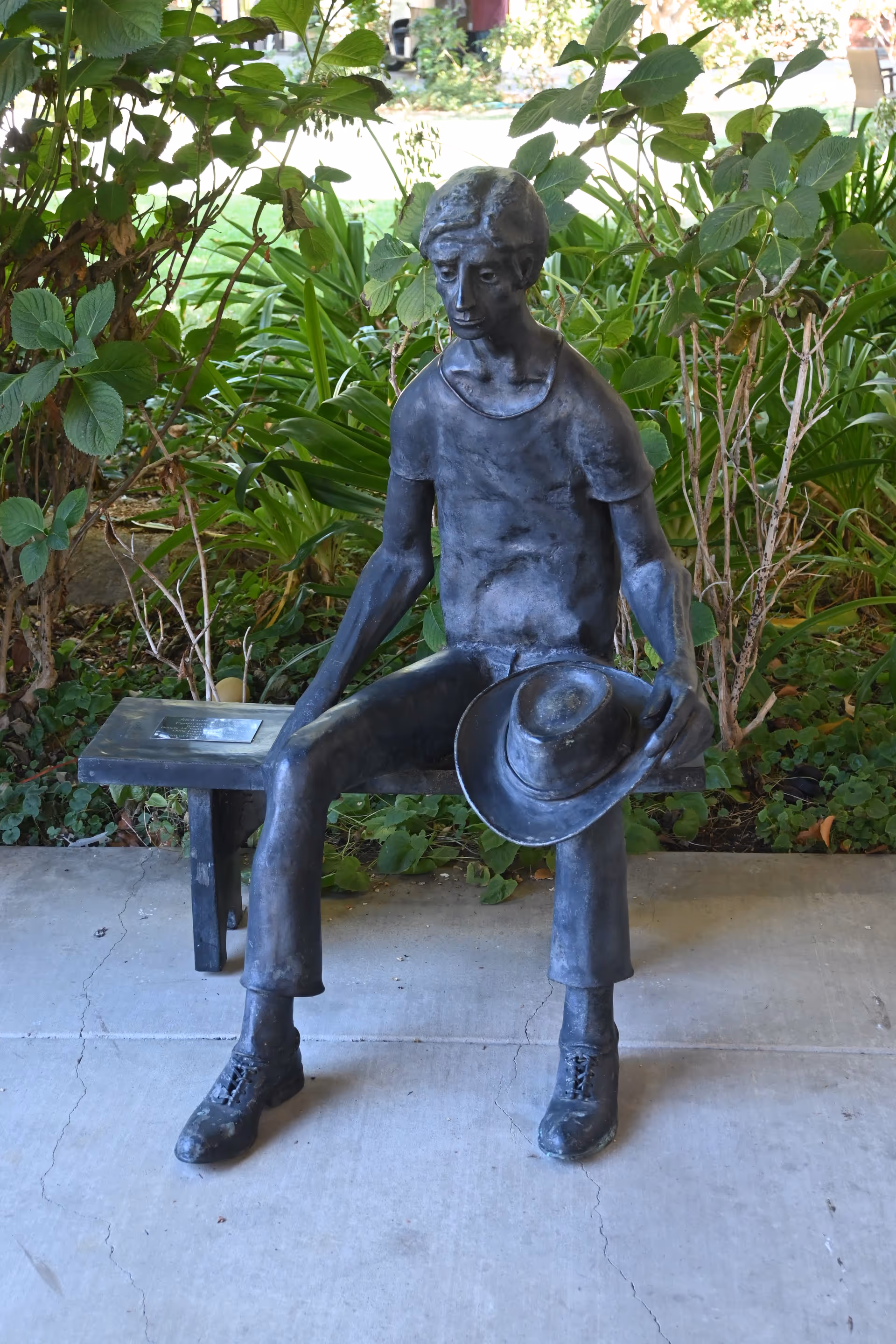 Bronze statue of a seated man holding a hat in a shaded garden.
