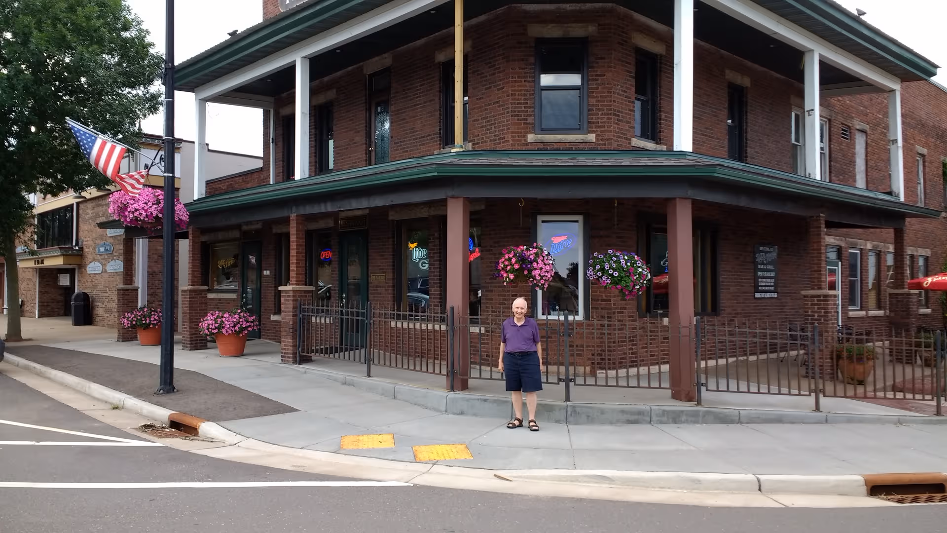 Brick senior living building on a corner with hanging baskets and a person standing outside.