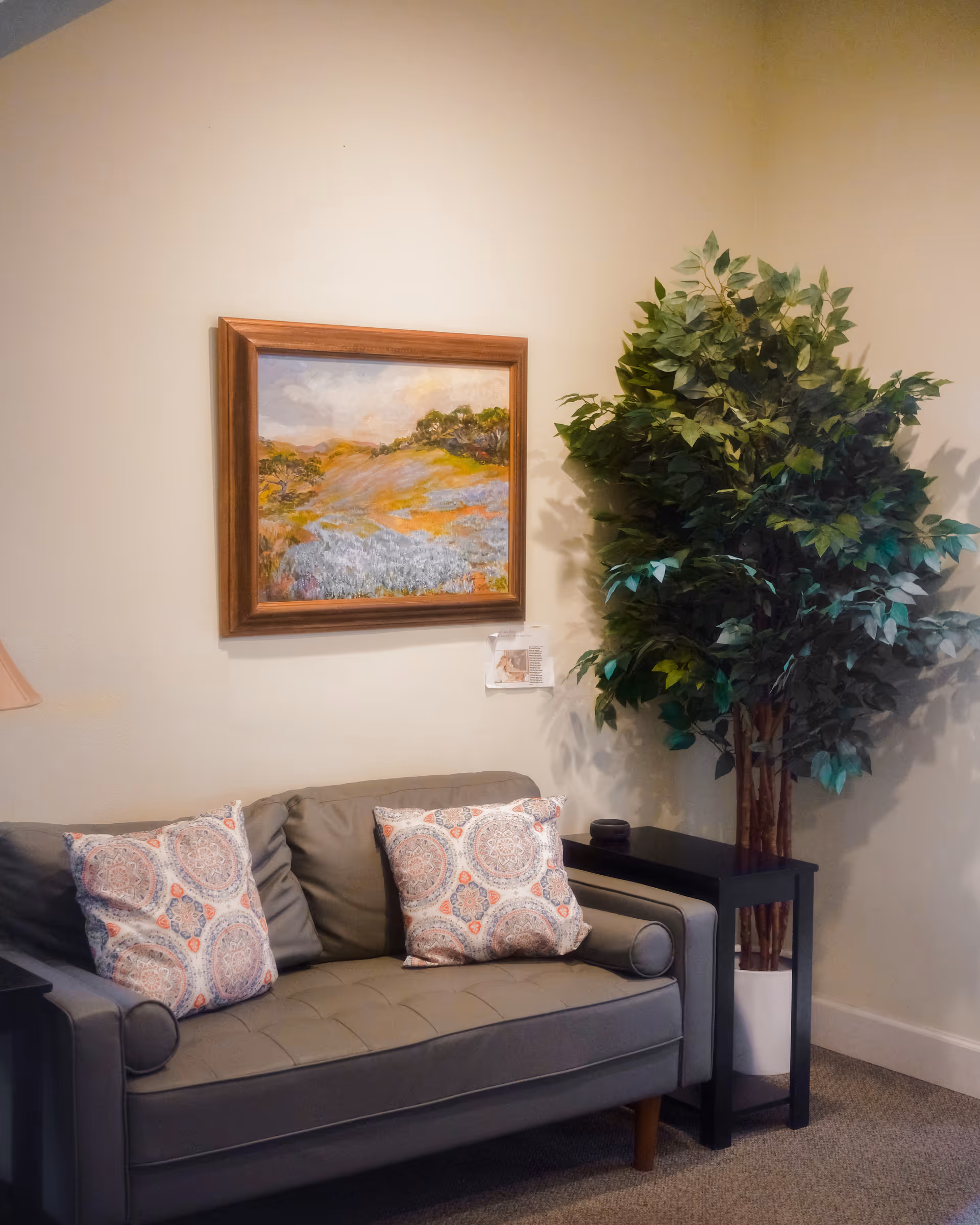 Cozy living room seating area with a gray sofa, decorative pillows, a painting, and a plant.