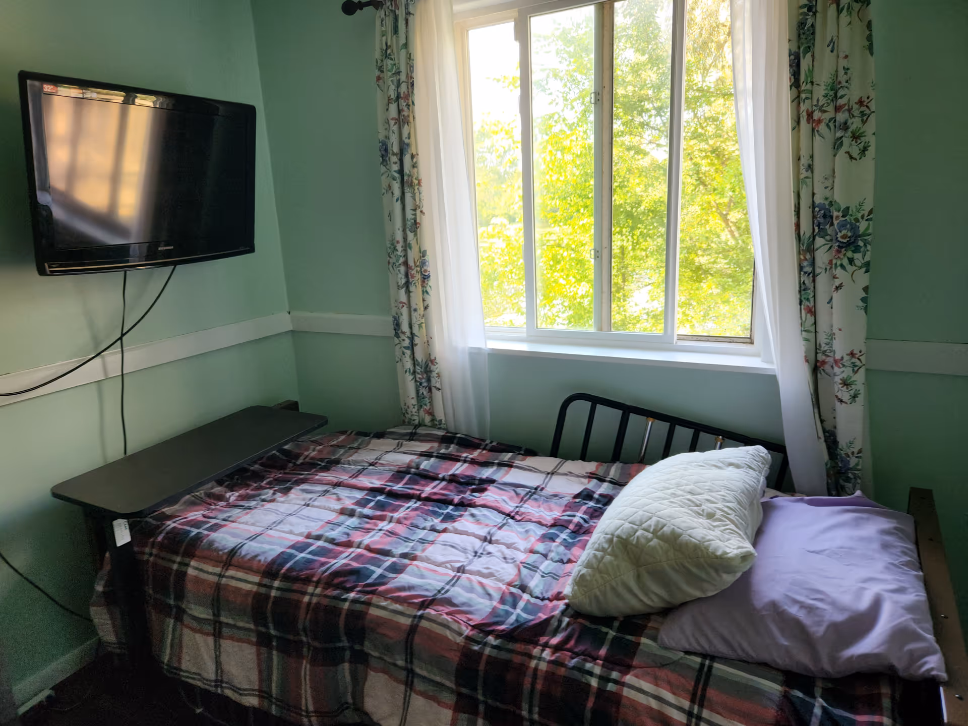 Small bedroom with single bed, plaid quilt, wall-mounted TV, and sunny window.