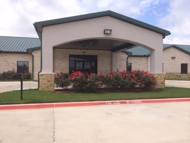 Front entrance of a single-story senior living facility with a covered porte-cochère, flowering bushes, and a green metal roof.