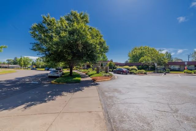 Parking lot area with several parked cars and a large tree in the center island. The building of Corvallis Manor Nursing & Rehabilitation Center is visible in the background under a clear blue sky.