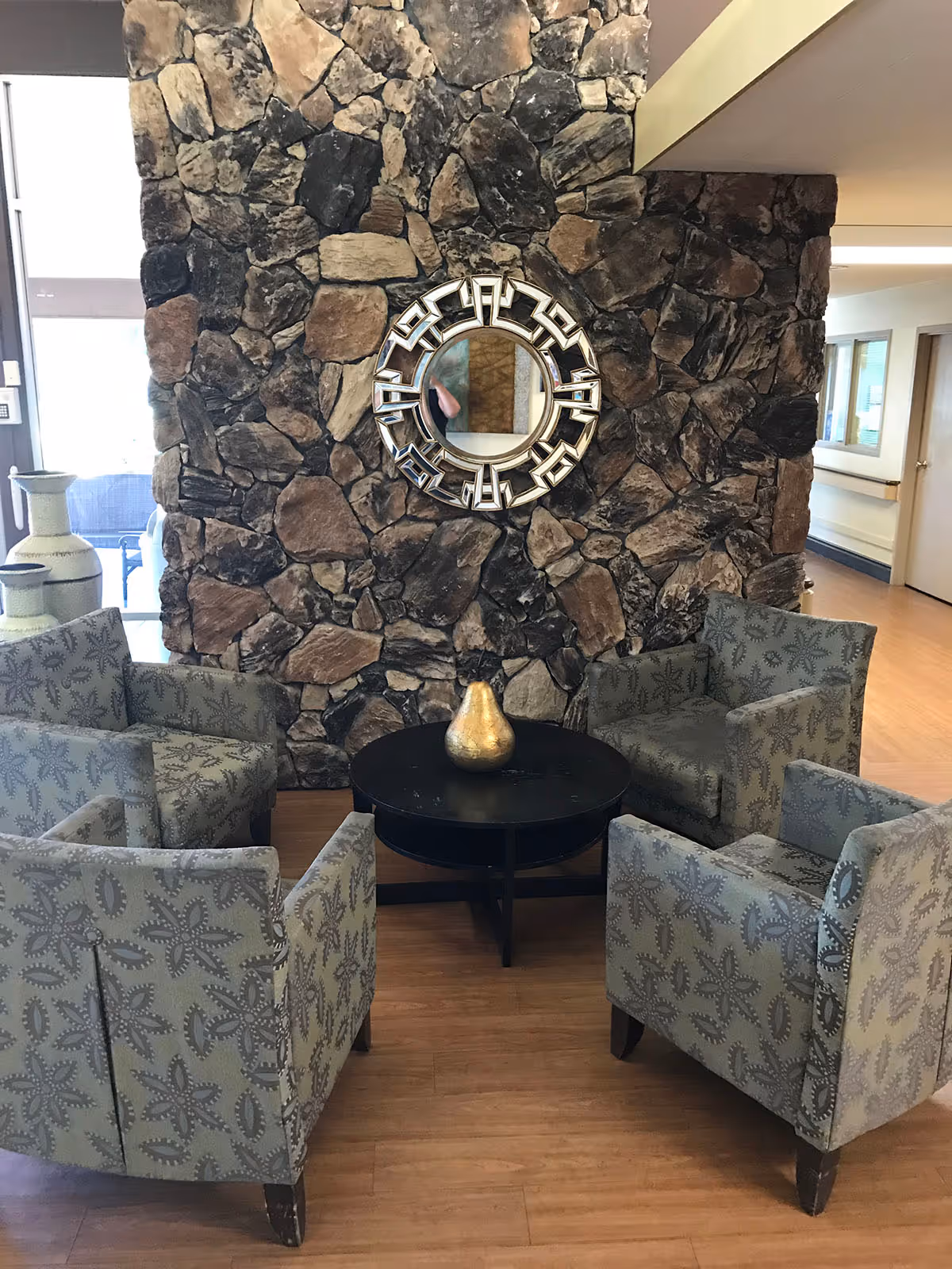 Seating area with four patterned armchairs around a round black table in front of a stone accent wall with a decorative round mirror.
