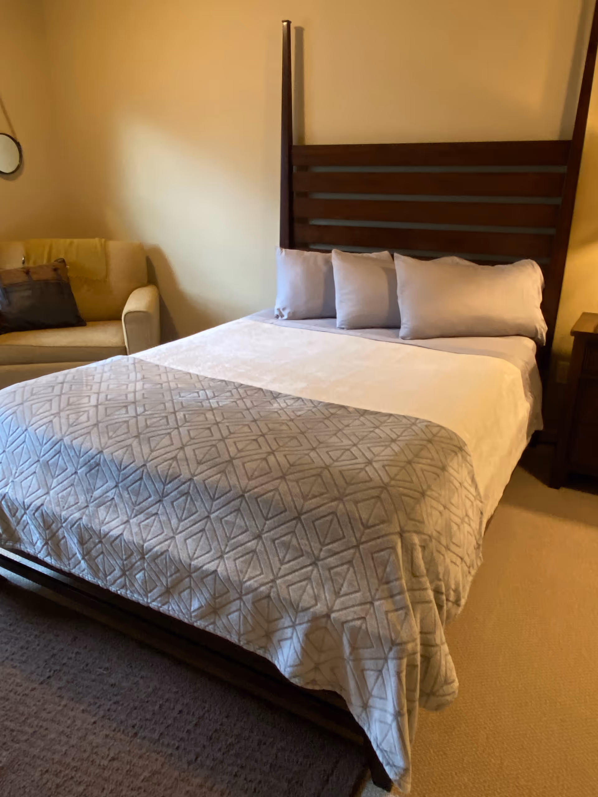 A neatly made bedroom with a dark wooden headboard, pillows, a sofa, and a nightstand.