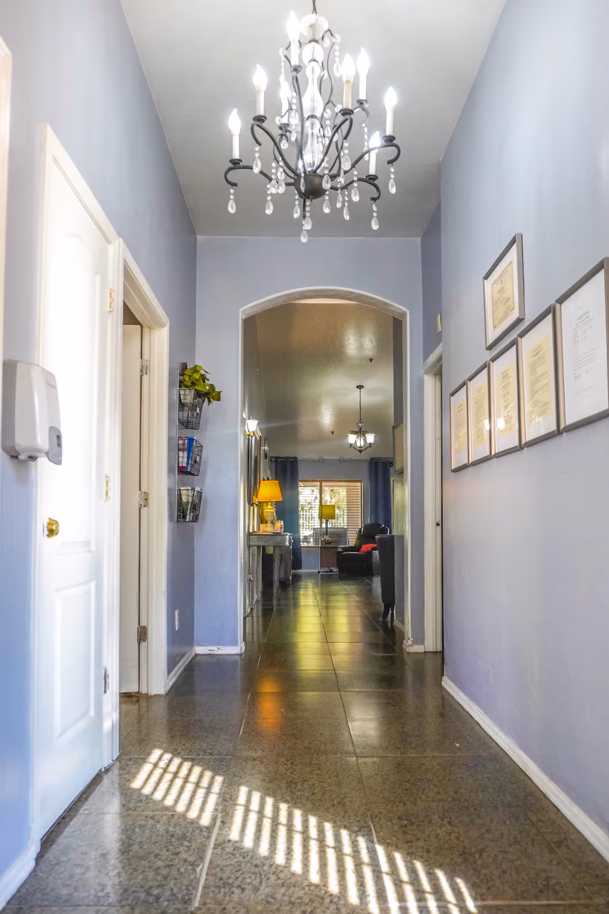 A lavender-walled hallway with chandelier, archway, and framed documents in a senior living facility.
