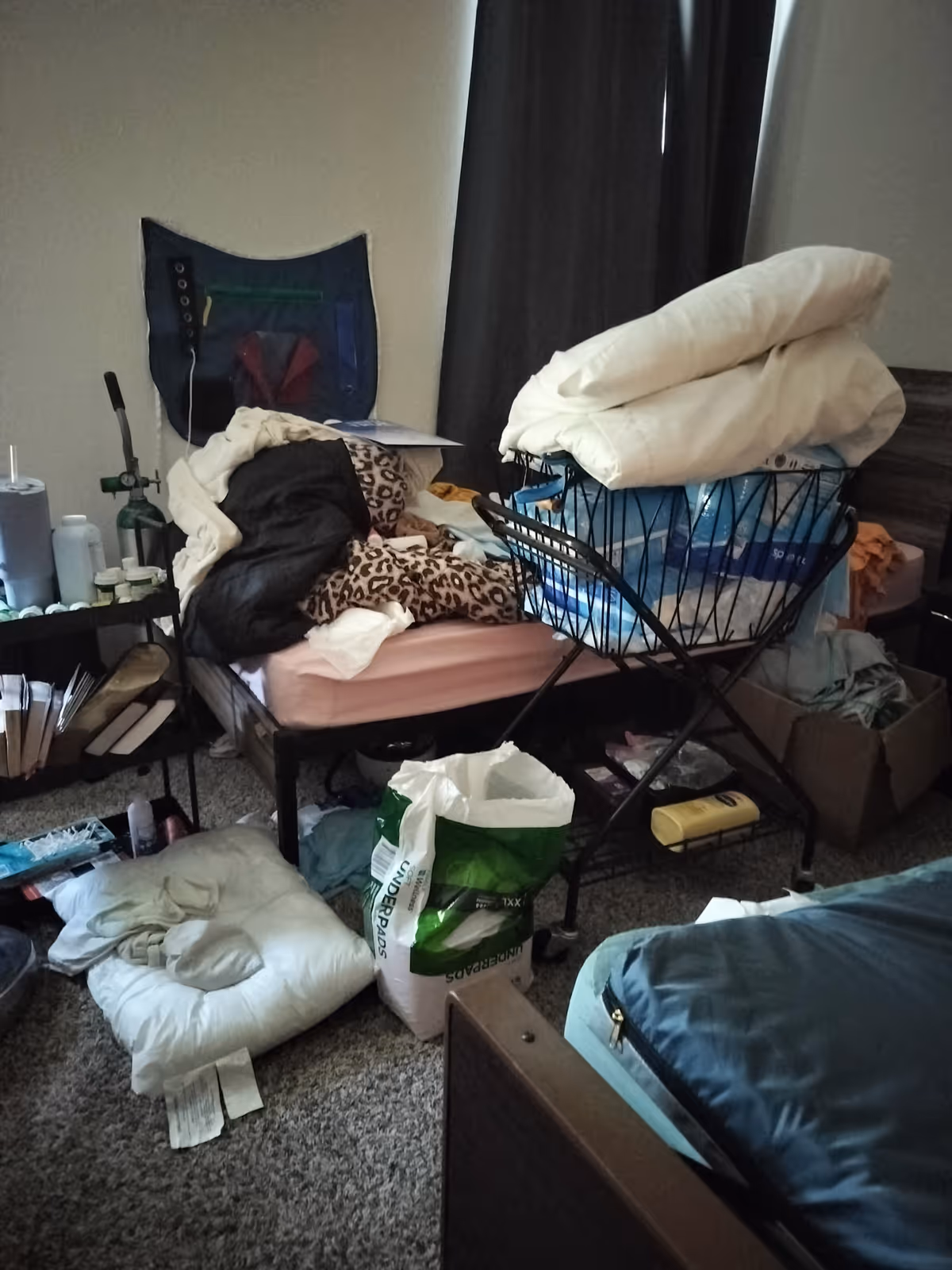 A cluttered bedroom in a senior living facility with a bed, laundry pile, and scattered items.