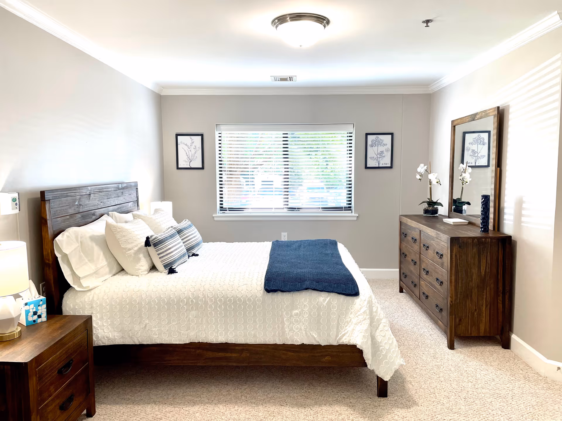 A neatly made bedroom with a wooden bed, dresser, nightstands, and window blinds.