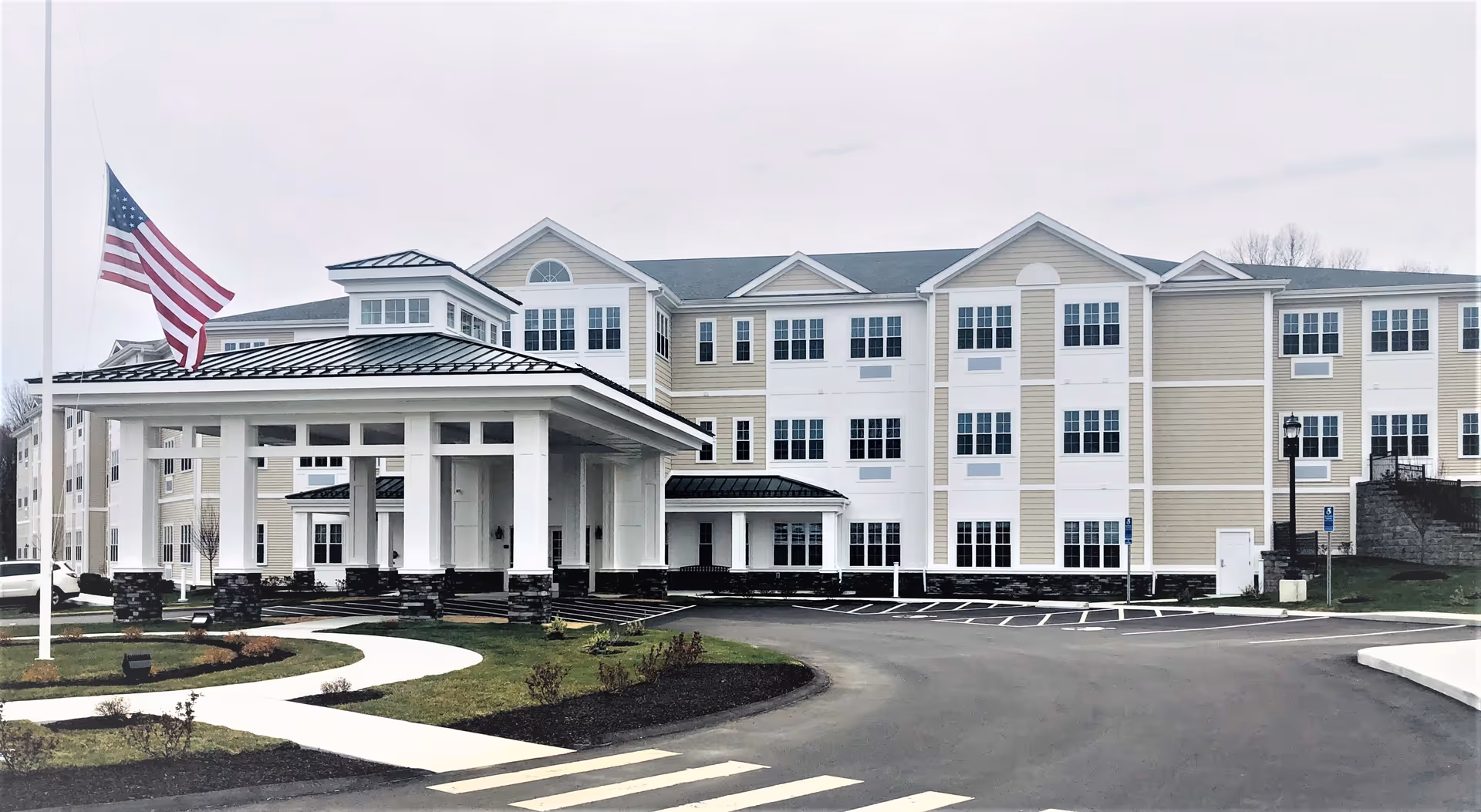 Exterior view of a large, multi-story assisted living facility building with beige and white siding, numerous windows, and a covered entrance with white columns and a black roof. An American flag is flying at half-mast near the entrance. There is a paved driveway and landscaped grass areas with small bushes in front of the building.