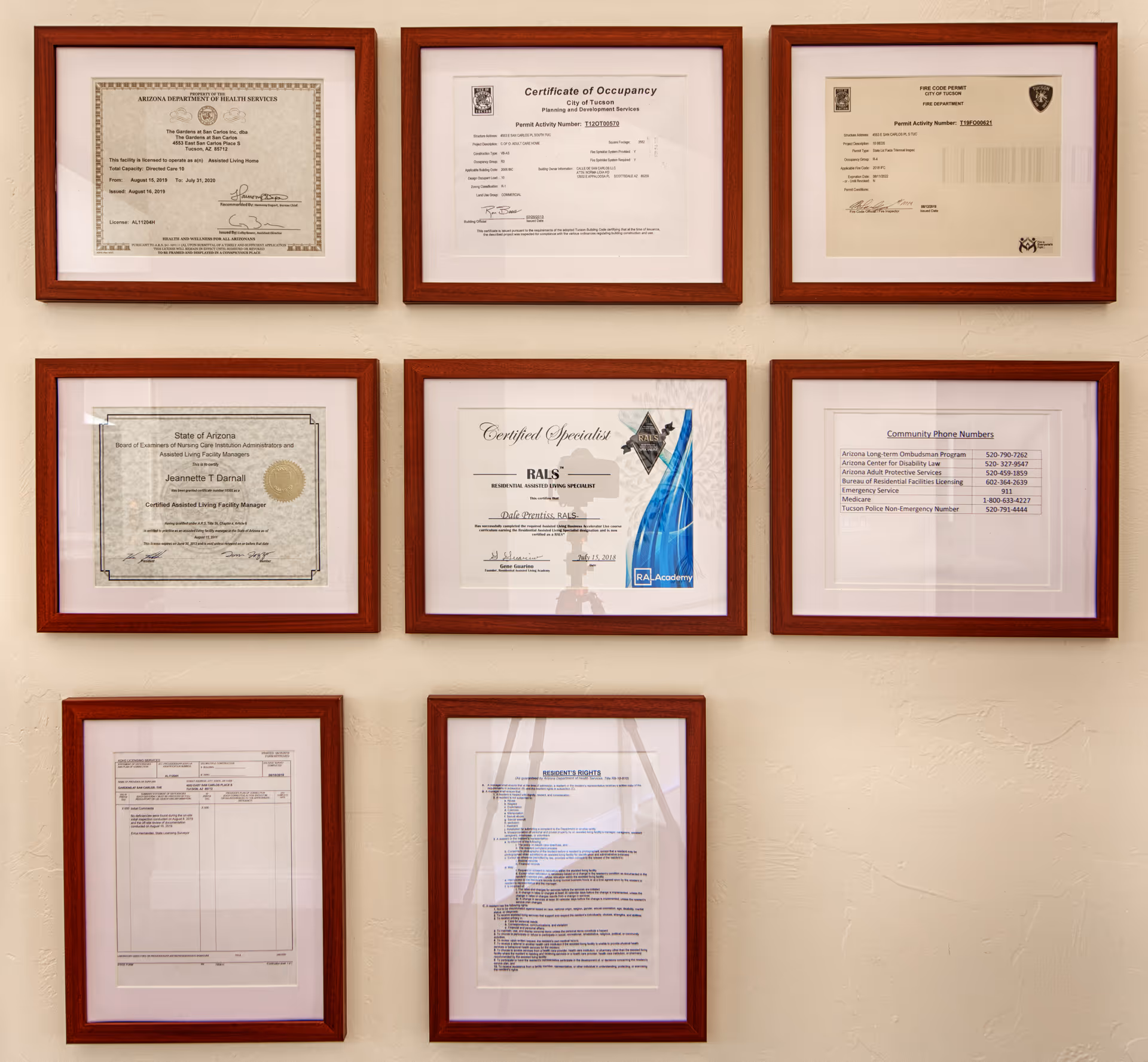 A wall displaying seven framed certificates and documents related to The Gardens at San Carlos, including licenses, permits, specialist certifications, community phone numbers, and resident rights.