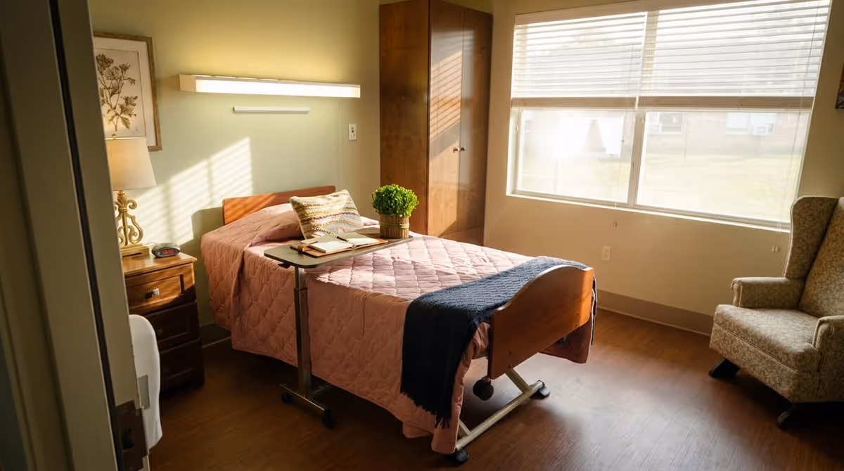 A furnished patient bedroom with a pink quilt bed, window, and chair.