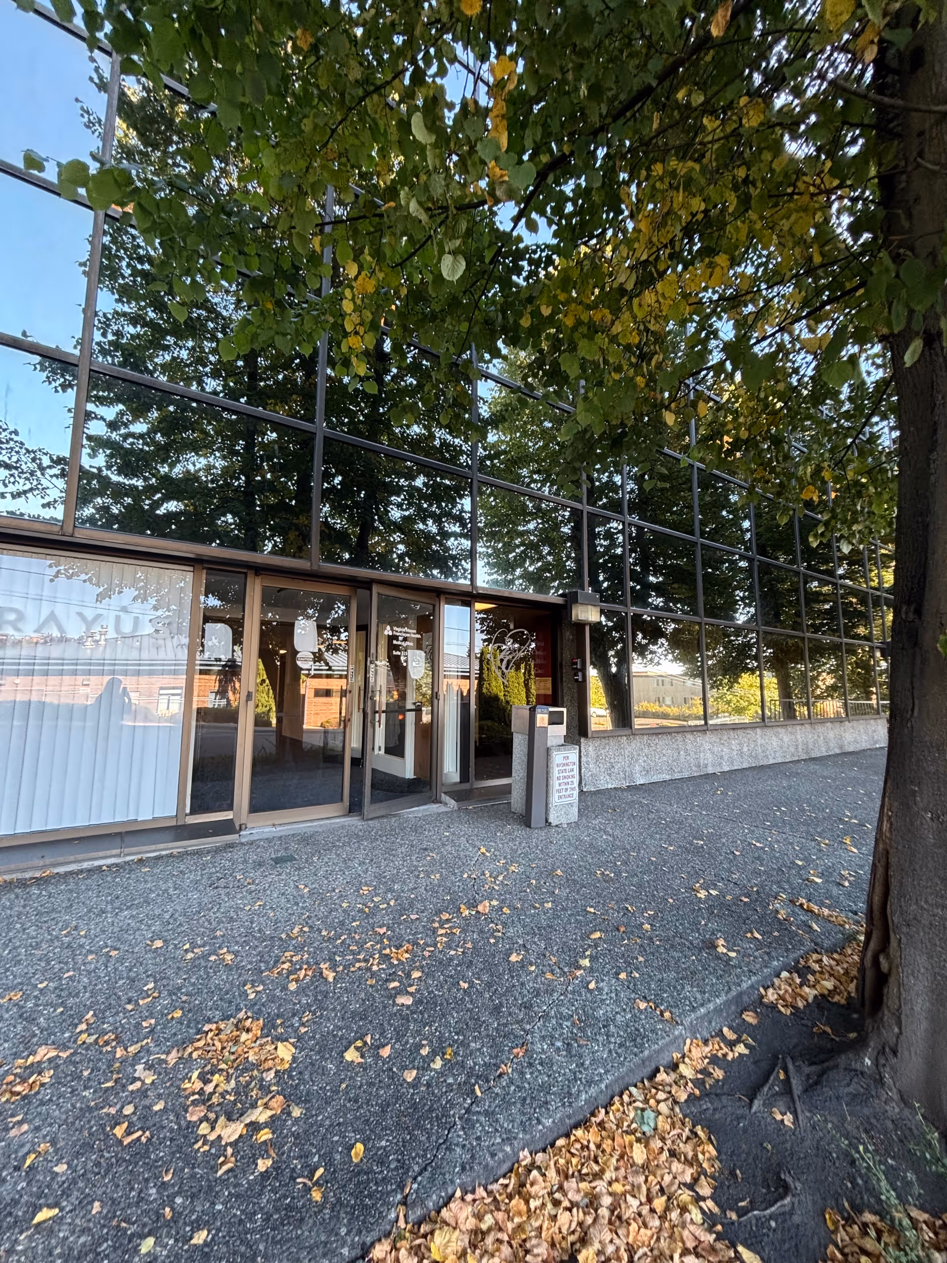 Glass-front building entrance with trees, autumn leaves, and a paved walkway.