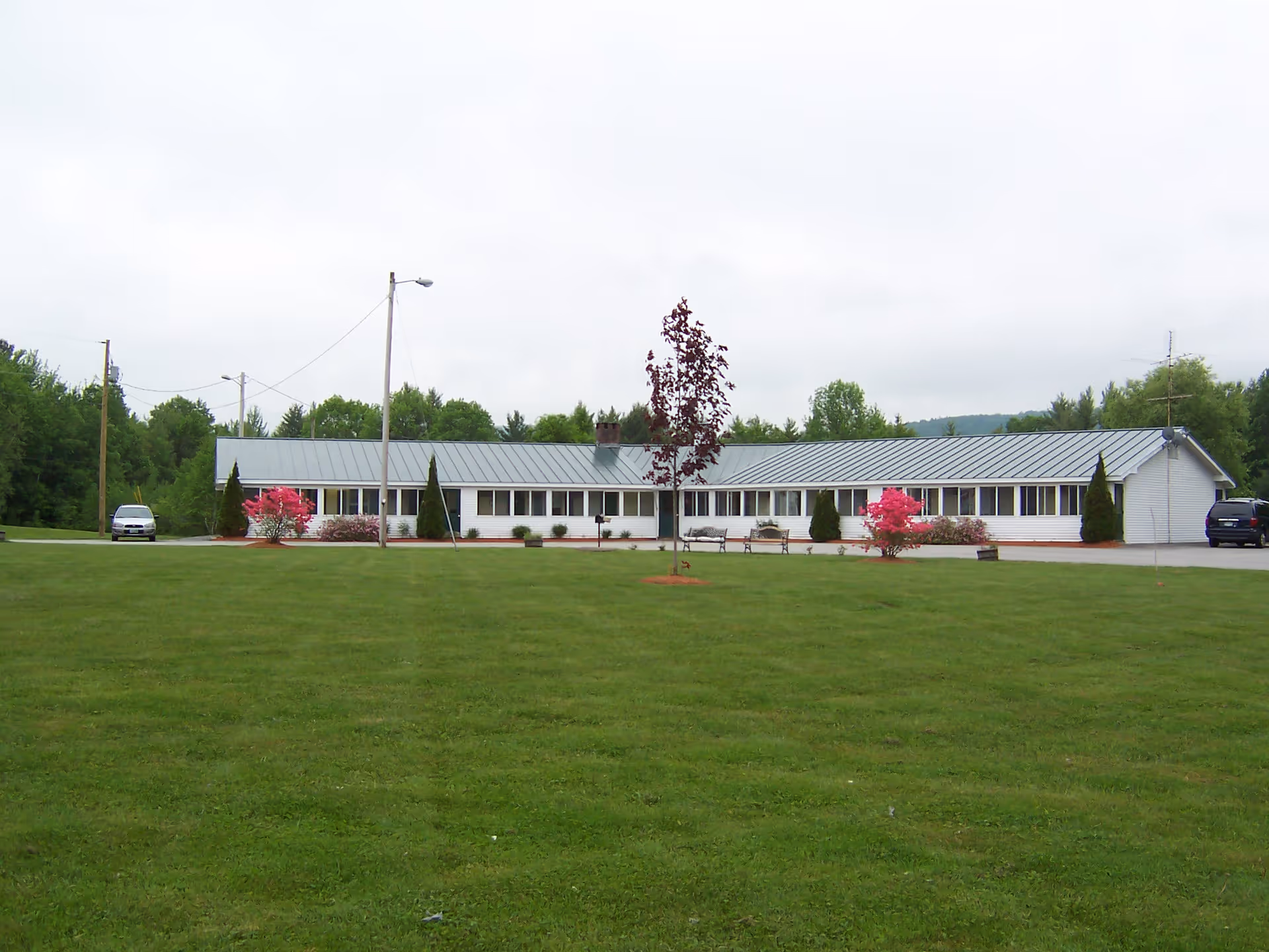 A single-story white building with a metal roof situated behind a large, well-maintained grassy lawn. There are a few small trees and bushes with pink flowers in front of the building, along with two benches. Two cars are parked on either side of the building, and there are utility poles and trees in the background under a cloudy sky.