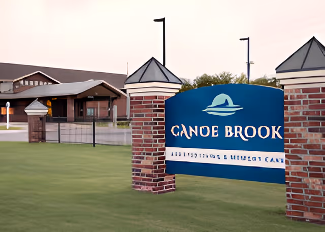Brick-pillared entrance sign reading "Canoe Brook" on a lawn with the assisted living facility building visible in the background.