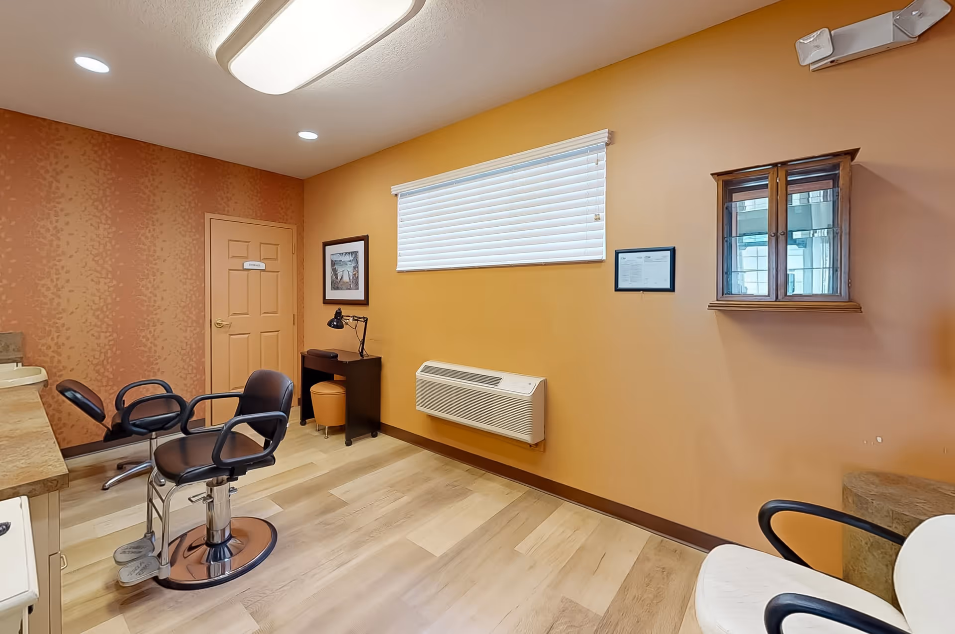 Interior room with two black salon chairs, a countertop with a sink, a small desk with a lamp and stool, a wall-mounted cabinet with glass doors, a window with closed blinds, and a door. The walls are painted in warm tones and the floor has light wood flooring.