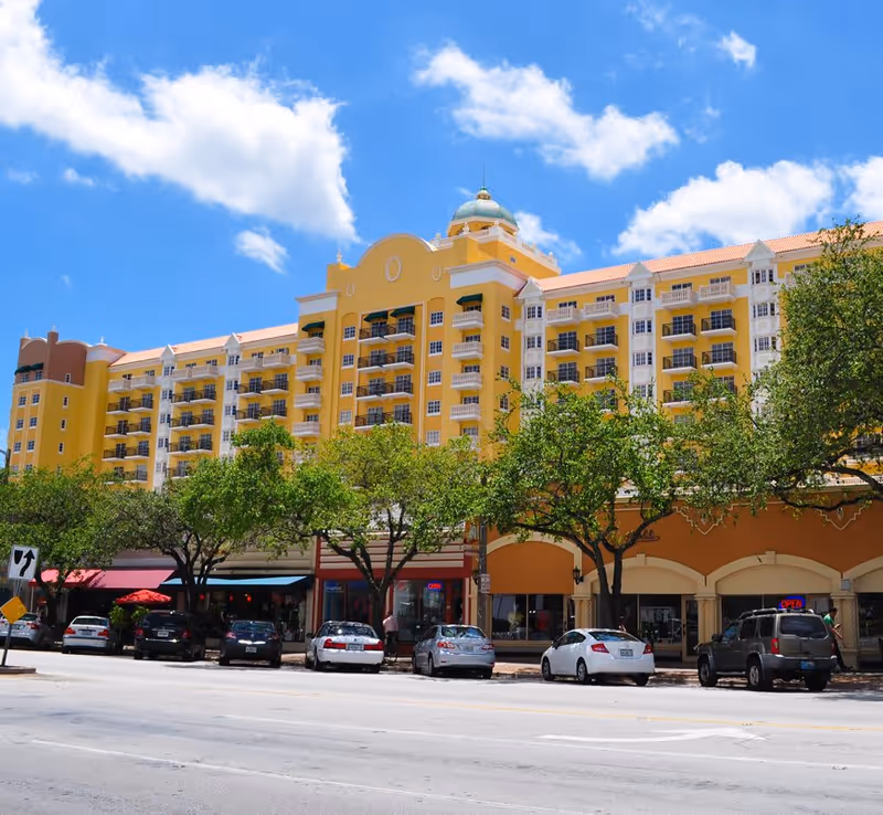 Front view of The Palace at Coral Gables, a yellow multi-story building with balconies, street-level shops, trees and parked cars.