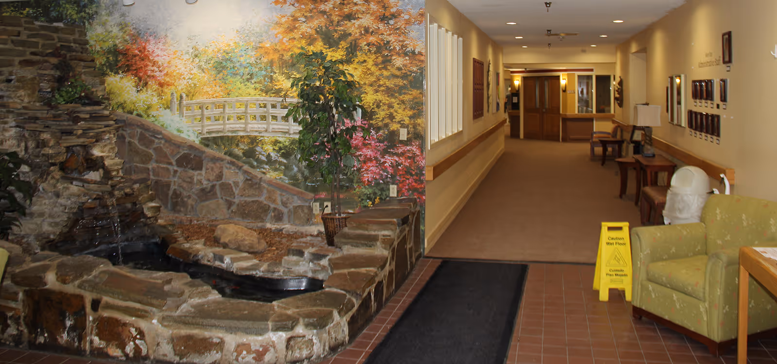 Interior hallway of Loyalhanna Care Center with a stone water feature and mural on the left, seating and a caution wet floor sign on the right, leading to doors at the end.