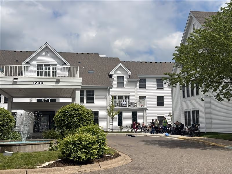White senior living facility exterior with entrance, fountain, and residents gathered outside.