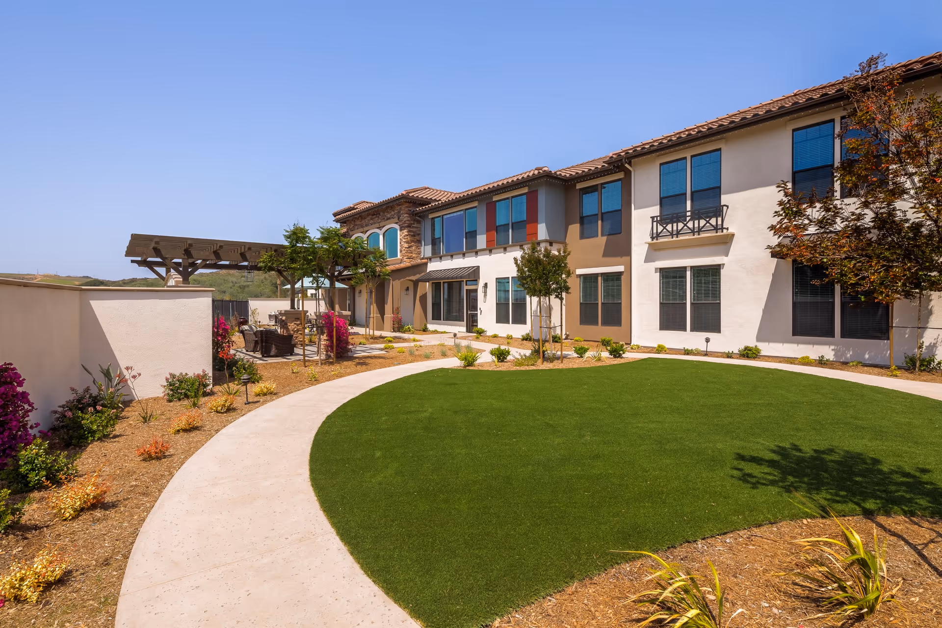 Courtyard with a circular lawn and walkway in front of a two-story memory care building with outdoor seating and a pergola.