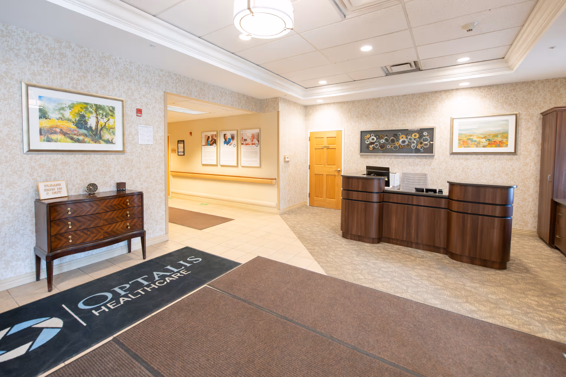 Reception area of Optalis Health & Rehabilitation of Canton (MI) featuring a wooden reception desk with a computer, a wooden chest of drawers with a small sign, framed artwork on the walls, and a floor mat with the Optalis Healthcare logo.