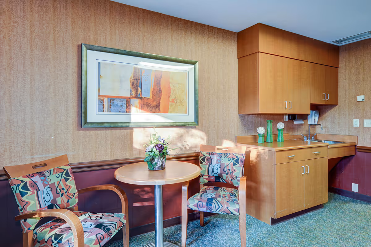 A small lounge area with a round table, patterned chairs, wall art, and a kitchenette.