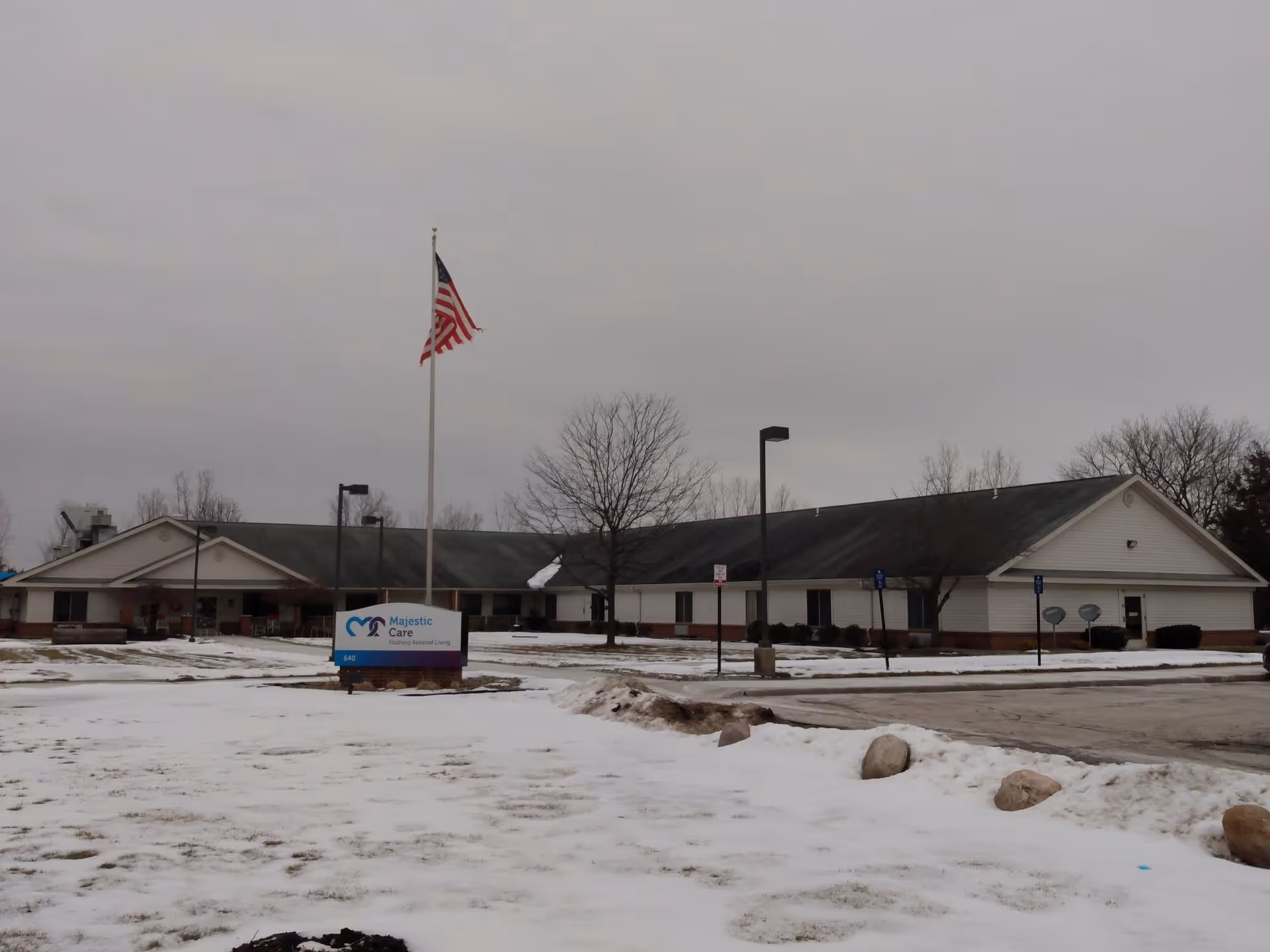 Exterior view of Majestic Care of Flushing Assisted Living facility on a cloudy day with snow covering the ground. The building is single-story with a pitched roof, and there is an American flag on a flagpole in front of the building. A sign with the facility's name is visible near the flagpole.