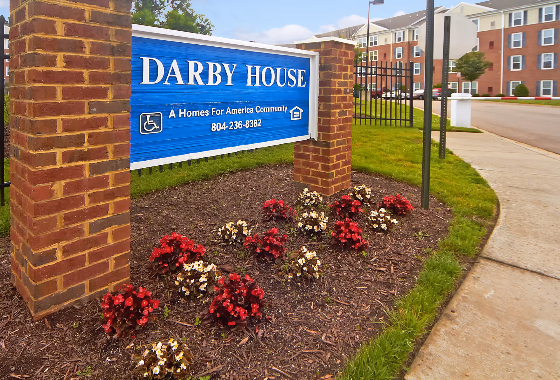 Entrance sign for Darby House, a Homes For America community, mounted on a blue board supported by two brick pillars with red and white flowers planted in front. In the background, there is a sidewalk, a street, and a multi-story residential building.