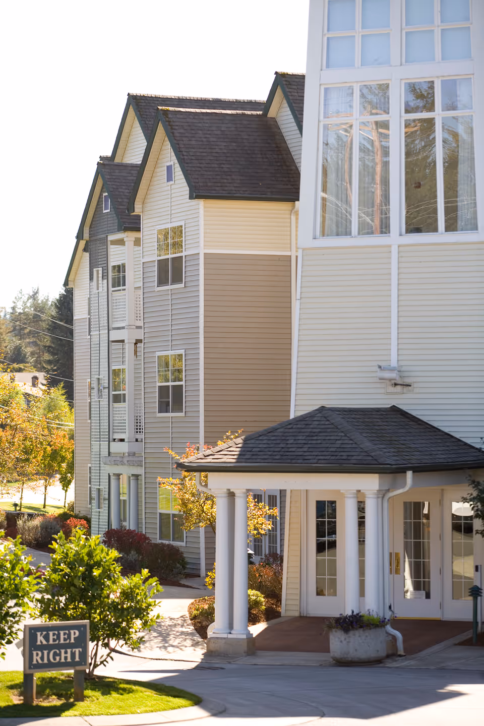 Exterior view of a multi-story senior living facility with beige and gray siding, white columns supporting a covered entrance, and large windows. There is a 'KEEP RIGHT' sign on a grassy area near the driveway, with trees and shrubs surrounding the building.