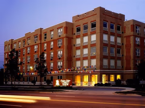 Exterior view of a brick senior living building with lit entrance and bunting at dusk.
