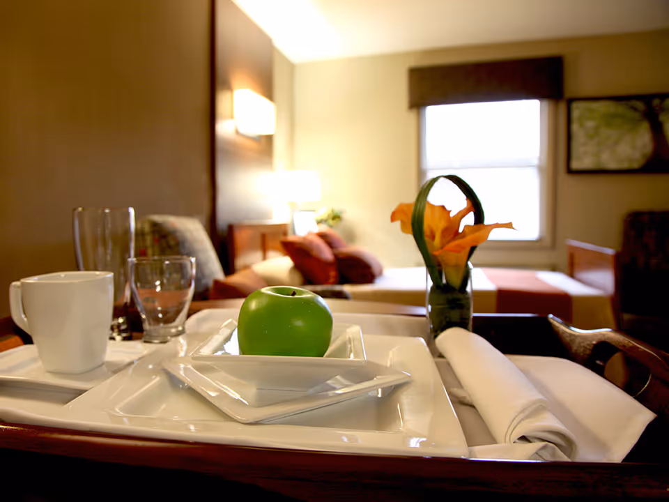 A breakfast tray with white dishes, a green apple and a vase of orange flowers on a table in front of a bedroom with a bed and window.