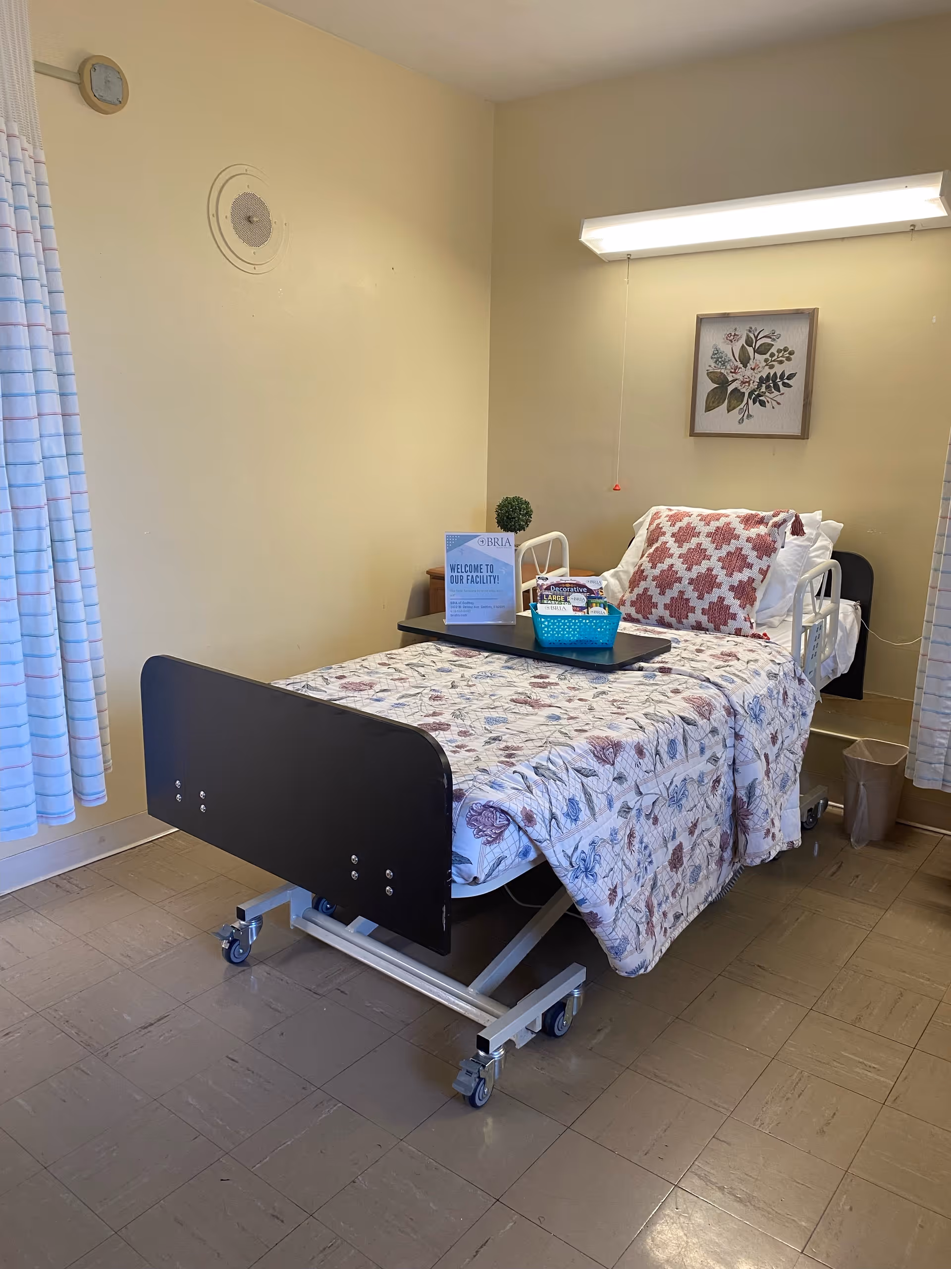 A single hospital-style bed with floral bedding and a red and white patterned pillow in a small room. A tray table on the bed holds a welcome sign, a small green plant, and a basket with snacks. The room has beige walls, a framed floral picture above the bed, a trash can in the corner, and a window with striped curtains.