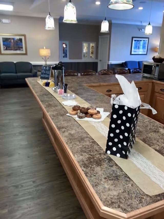 Interior view of a senior living facility common area with a large kitchen island countertop. On the countertop, there is a black and white polka dot gift bag with white tissue paper, a plate of assorted donuts, a coffee dispenser, and some condiments. In the background, there are chairs, a microwave, a couch, framed pictures on the walls, and hanging pendant lights above the island.