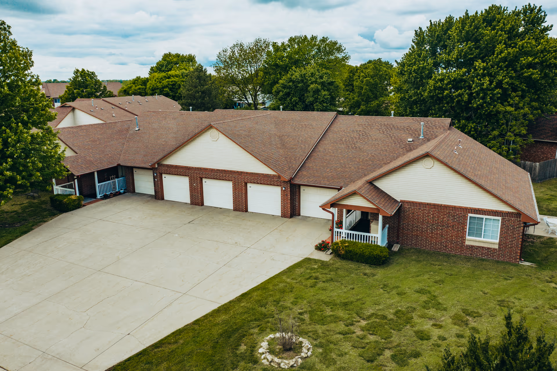 Aerial view of a single-story brick building with multiple garage doors and a small covered porch, surrounded by green grass and trees under a cloudy sky.