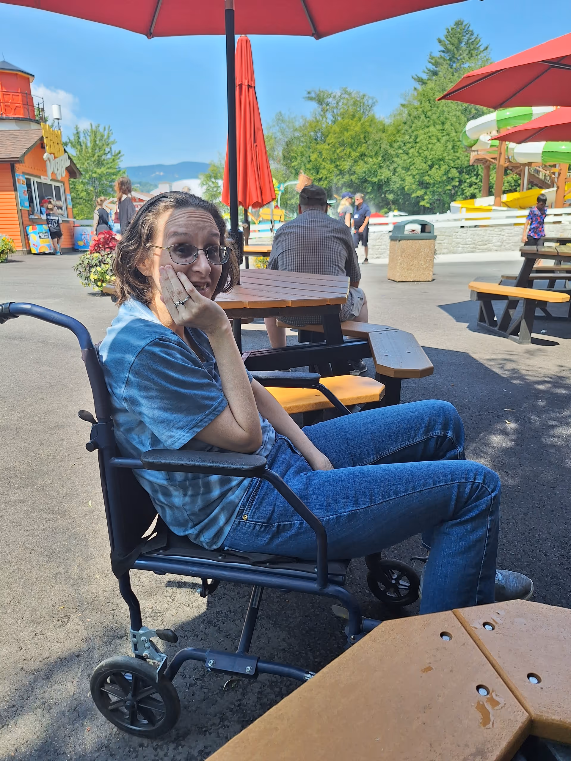 Woman in a wheelchair sits at an outdoor seating area with umbrellas.