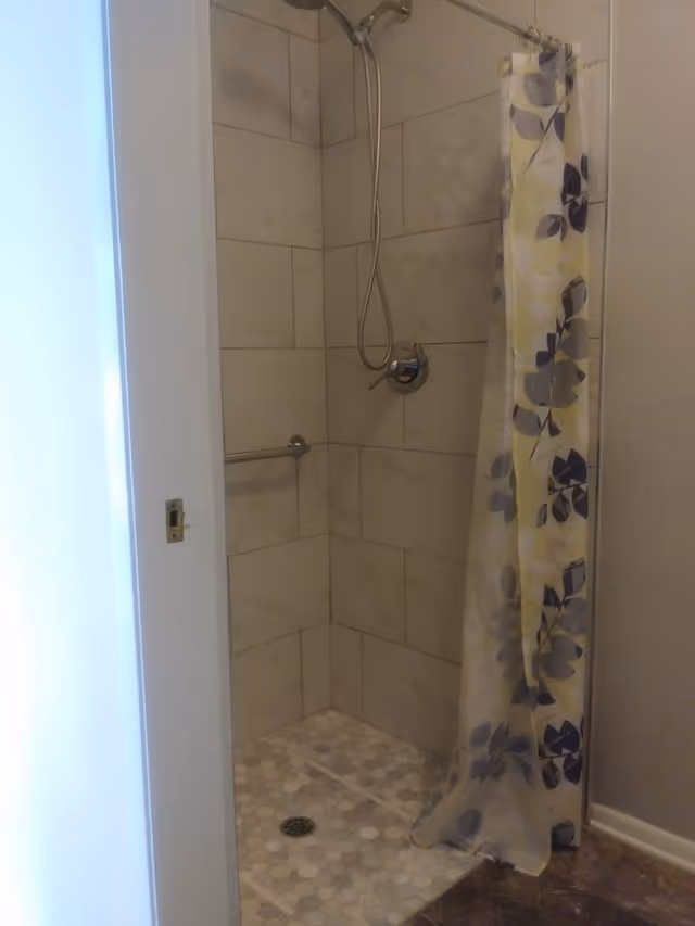 A tiled shower area with a handheld showerhead mounted on the wall, a metal grab bar, and a shower curtain with a floral pattern in shades of gray and purple. The shower floor has small hexagonal tiles in various shades of gray and white.