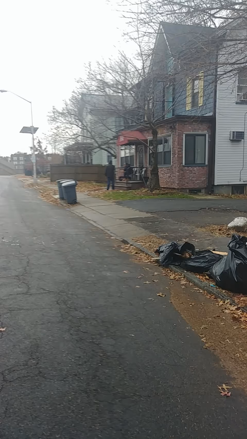 Residential street view with brick building, red awning, leaf litter, and trash bags.