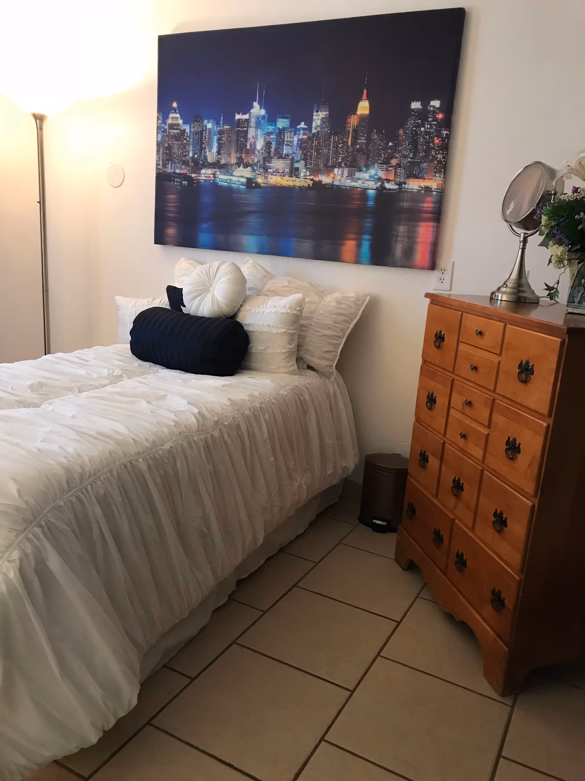A neatly made bed with white bedding, a dresser, and skyline artwork.