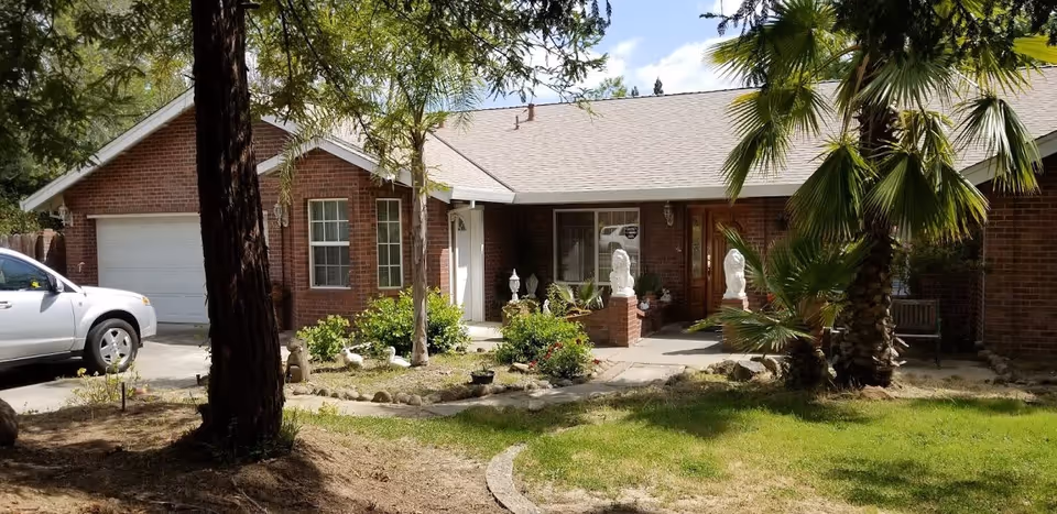 Front exterior of a single-story brick residence with a garage, a white SUV in the driveway, and a small landscaped yard with palm trees and garden statues.