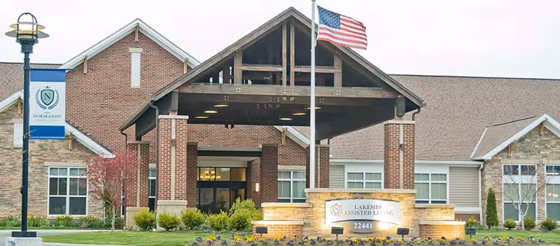 Front exterior view of Lakeside at the Normandy assisted living facility showing a covered entrance with brick pillars, an American flag on a flagpole, landscaped greenery, and a sign with the facility name and address.