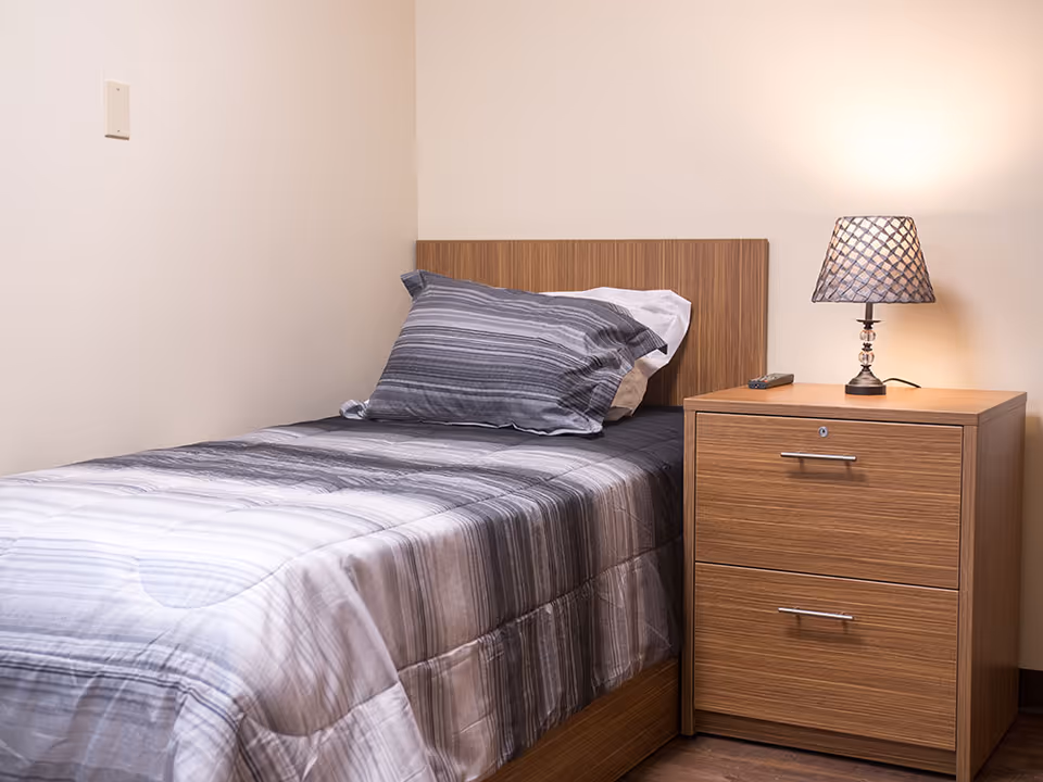 A single bedroom with a striped bed, wooden headboard, and a nightstand with lamp.