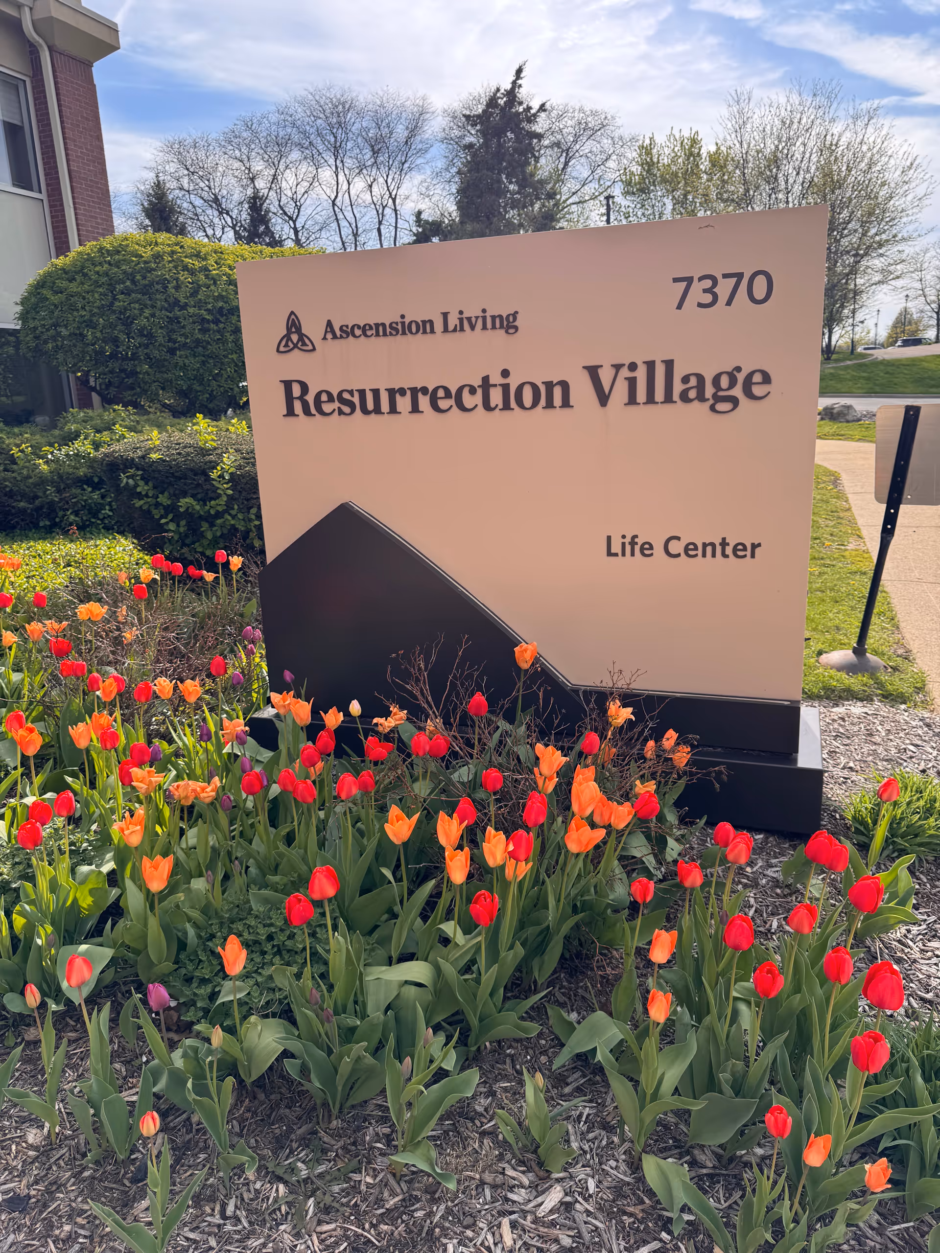 Outdoor sign reads Ascension Living Resurrection Village Life Center, surrounded by blooming tulips.