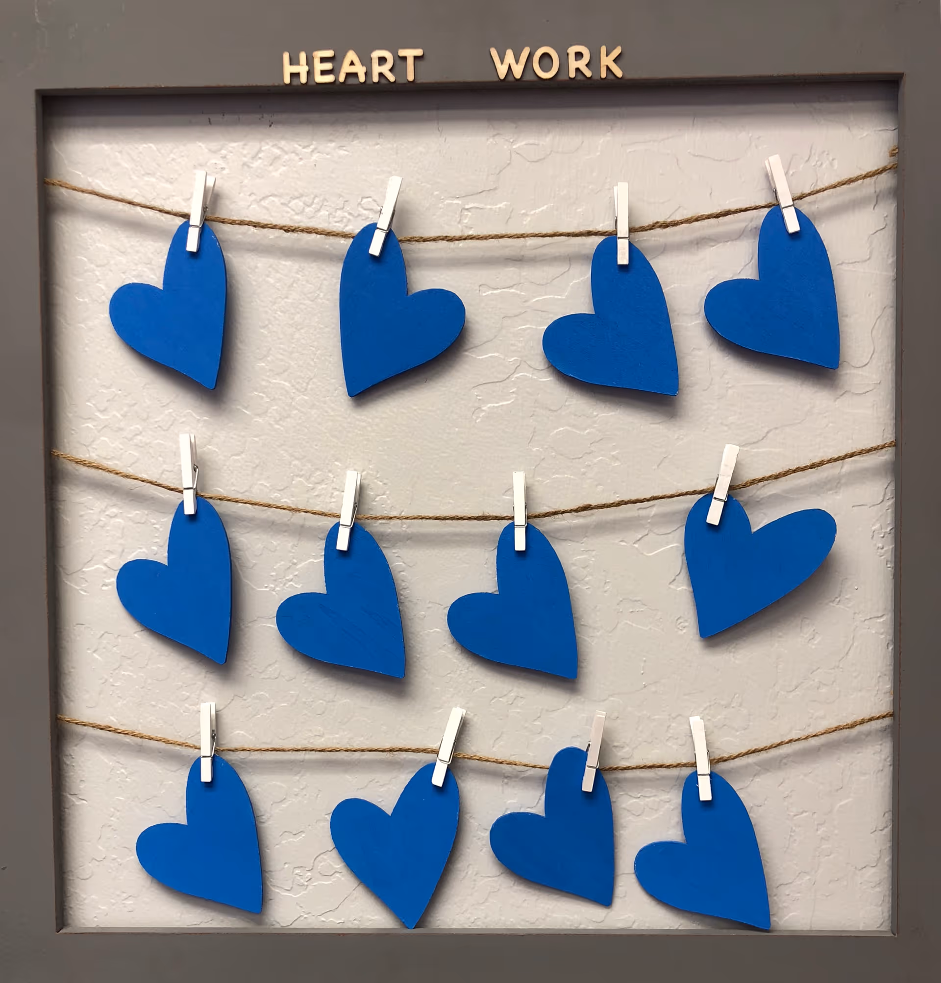 Framed wall display featuring blue heart shapes clipped to strings on a textured background.