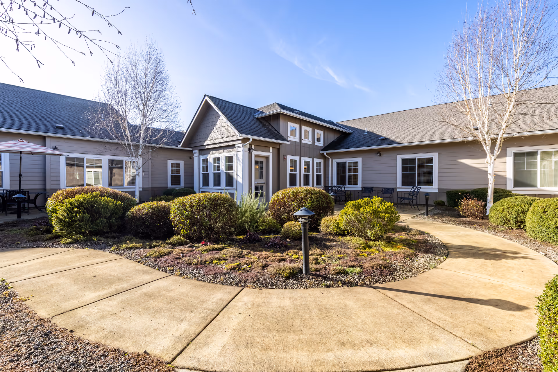 Exterior view of Heartwood Place Memory Care with curved walkway, landscaped courtyard, and outdoor seating.
