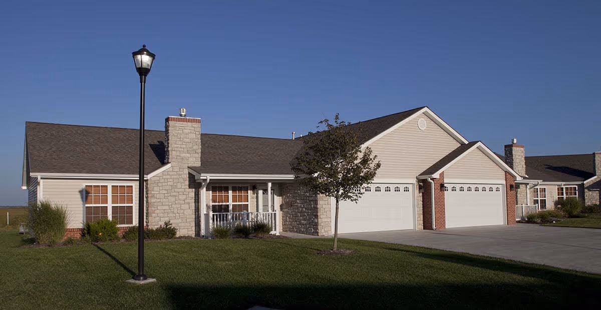 Front view of a single-story ranch-style building with three garage doors, a small porch, a lamp post, and a neatly mowed lawn under a clear blue sky.