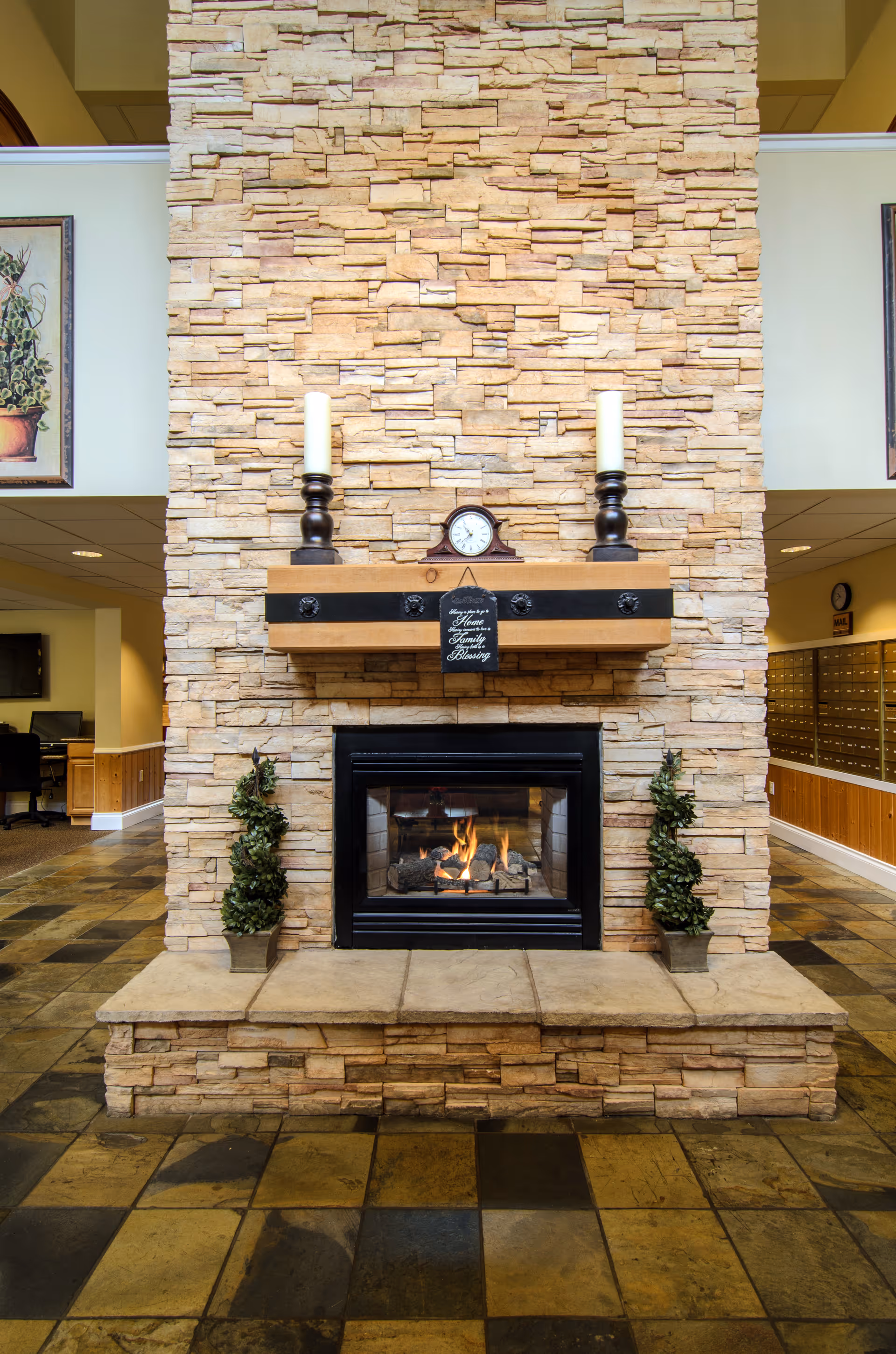 A stone fireplace with a wooden mantel holding two large candles and a small clock. Two small potted topiary plants flank the fireplace. The floor is tiled with a mix of brown and tan tiles. The background shows part of an interior space with mailboxes and office areas.