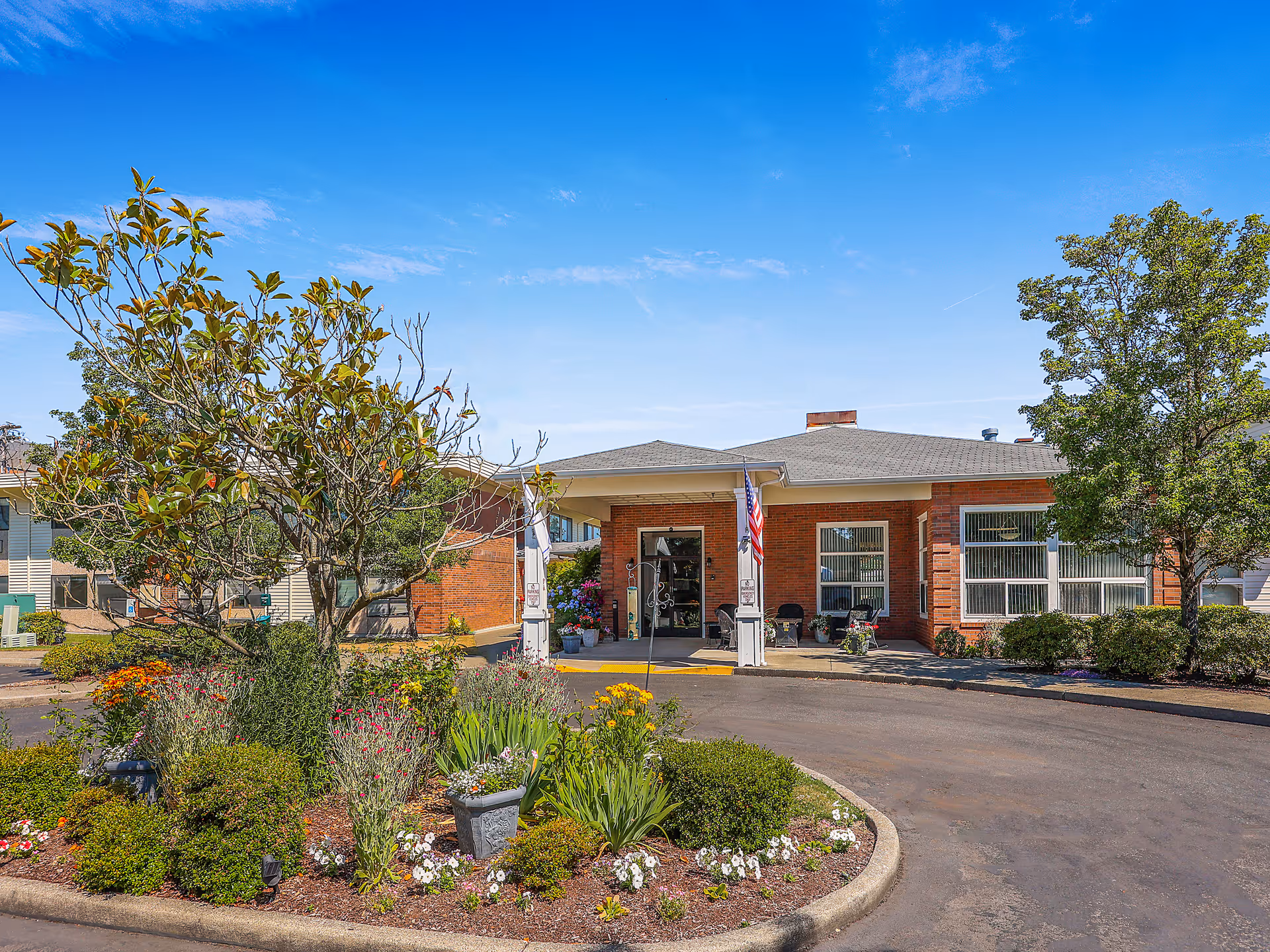 Front exterior view of a single-story brick building with a covered entrance, an American flag, and a landscaped circular driveway with various plants and flowers under a clear blue sky.